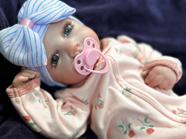 Lifelike reborn baby doll with blue eyes wearing a pink floral outfit and striped bow hat, realistic weighted newborn therapy doll
